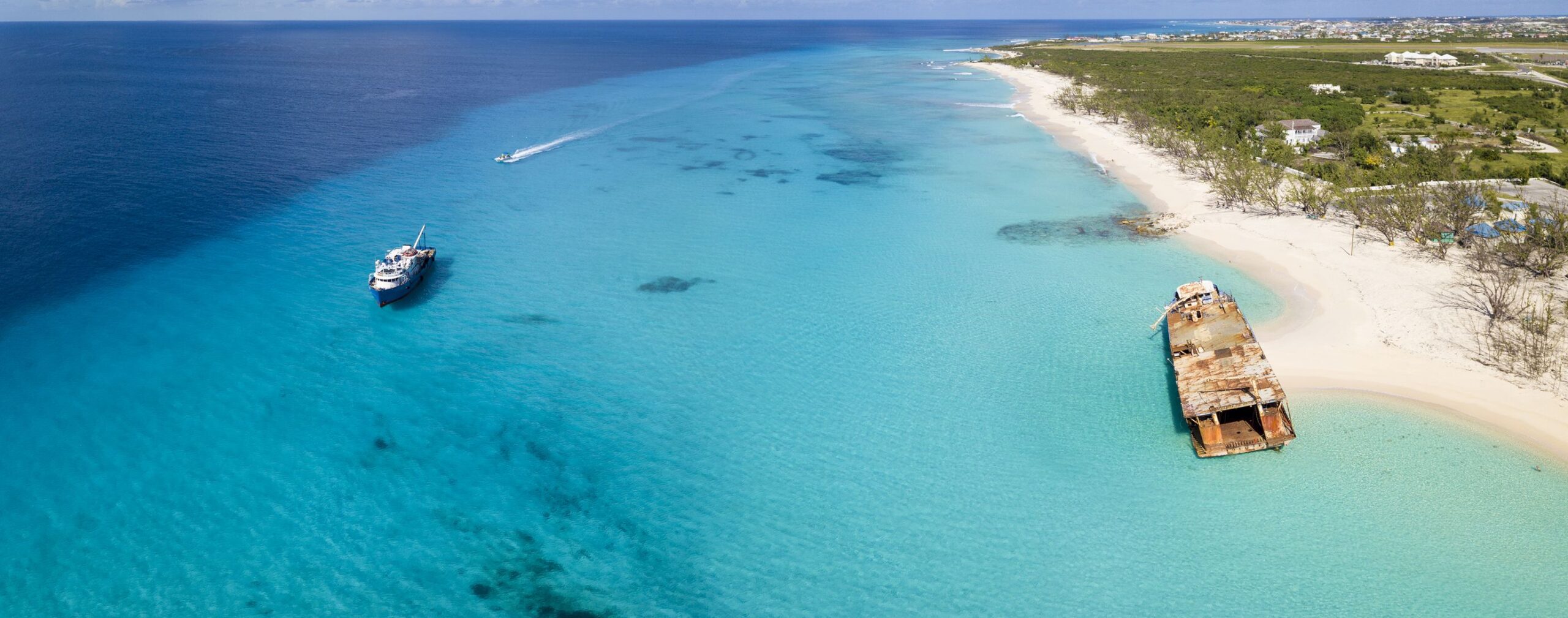 Aerial view of a tropical beach on the Turks and Caicos outer islands, with clear turquoise water, a rusted shipwreck near the shore, and a boat cruising nearby. Coastal greenery lines the sandy beach.