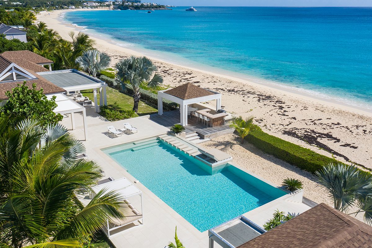 A luxurious beachfront villa with a large pool, sun loungers, shaded cabanas, and palm trees offers the privilege of breathtaking views as it overlooks a sandy beach and clear turquoise sea under a bright blue sky.