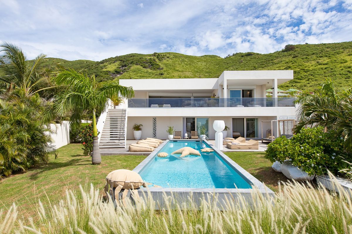 Modern white two-storey villa with large balconies, a swimming pool, lounge chairs, palm trees, and lush green hills in the background under a partly cloudy sky—an oasis of privilege and contemporary comfort.