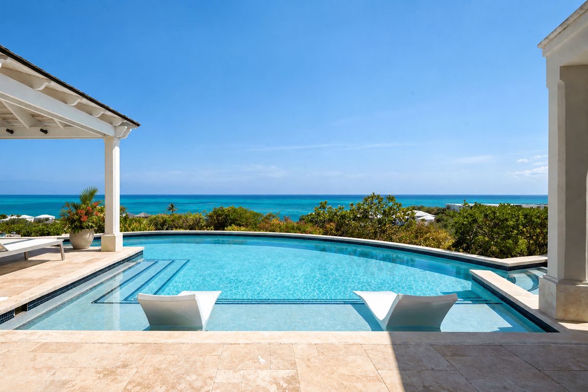 Infinity pool with two white sun loungers, stone patio, and a view of lush greenery leading to a turquoise sea under a clear blue sky. A covered terrace is on the left, and part of a white building frames the right.