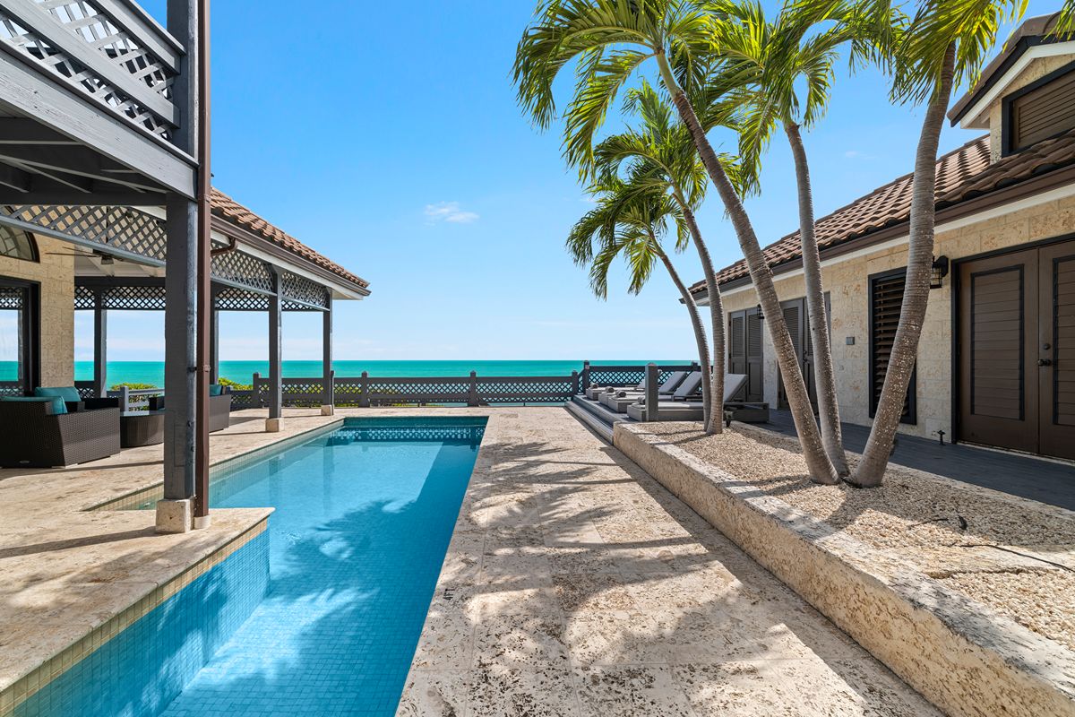 A swimming pool on the patio of the Taylor Bay Oceanfront Villa, overlooking the sea and framed by a covered terrace and palm trees.